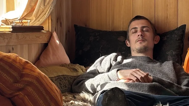 Filip hviler med lukkede øjne på en seng i et rum med træpaneler og varmt lys.